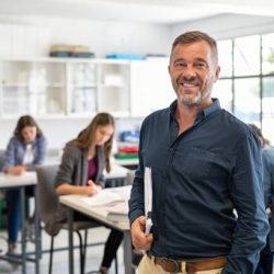 Portrait of mature teacher standing in university library and looking at camera with copy space. Happy mid adult lecturer at classroom standing after giving lecture. Satisfied high school teacher smiling and looking at camera while his studets studying in background.